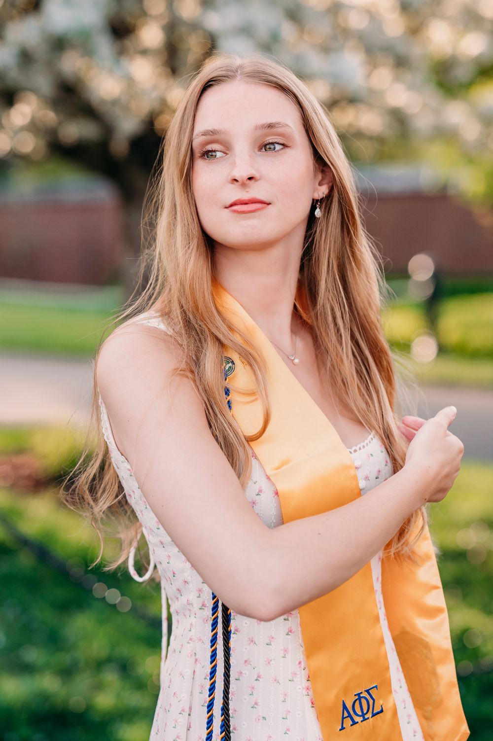 A person in a floral dress holds a yellow Alpha Phi Omega tote bag in a sunny outdoor setting.