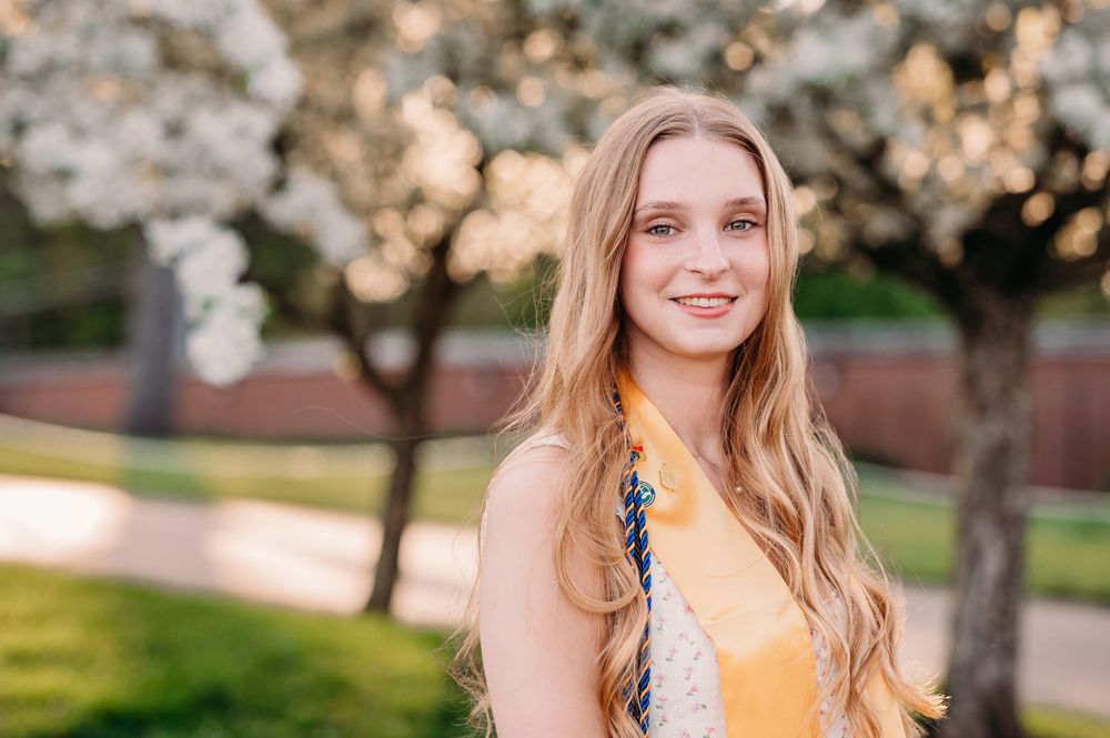 A person in a yellow dress stands outdoors with blooming trees and soft golden sunlight in the background.