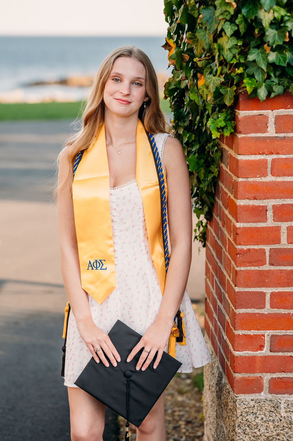 Graduate in white lace dress with yellow honor stole and diploma standing by brick wall with ivy.