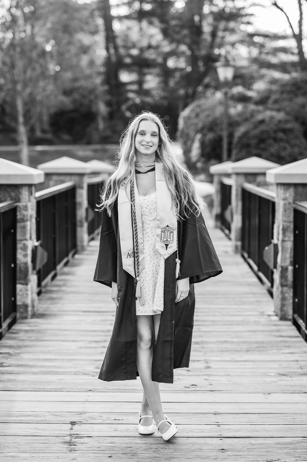 Black and white graduation photo showing graduate in cap and gown standing on a wooden bridge in an outdoor setting.