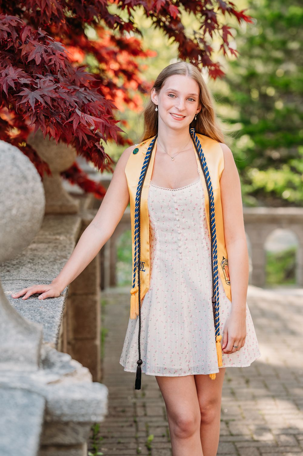 Graduate in white lace dress poses outdoors against vibrant red Japanese maple leaves and stone wall background.