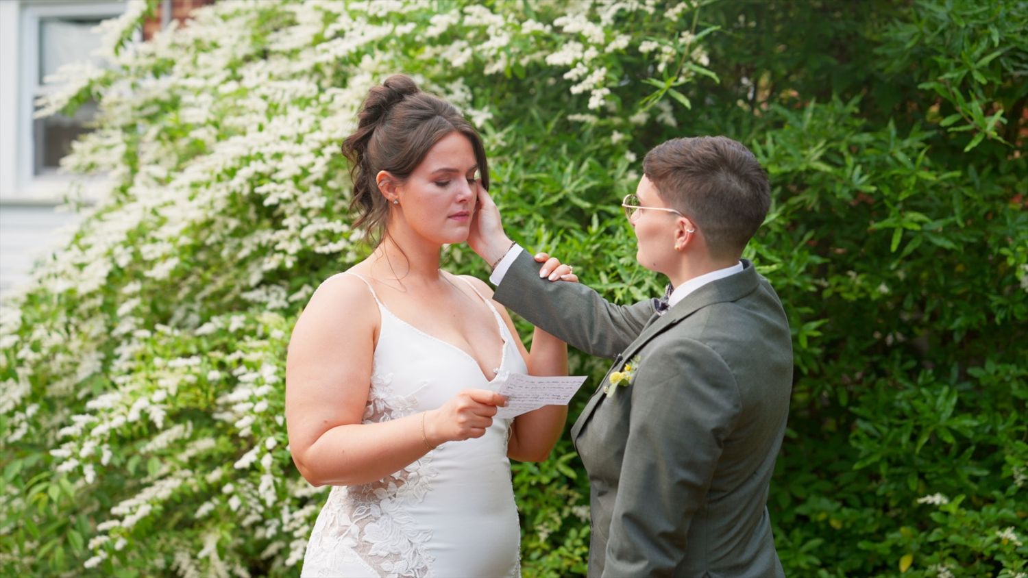 A bride and groom share an emotional moment outdoors during a wedding ceremony with greenery in the background.