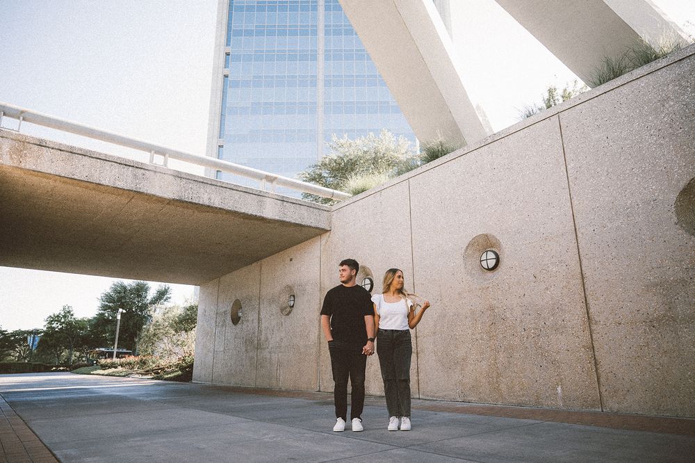 Two figures stand beneath a modern concrete overpass with a glass office building visible in the background.