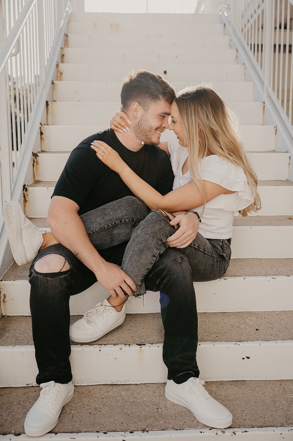 A couple sharing an intimate moment while sitting together on white concrete stairs dressed in casual black and white attire.