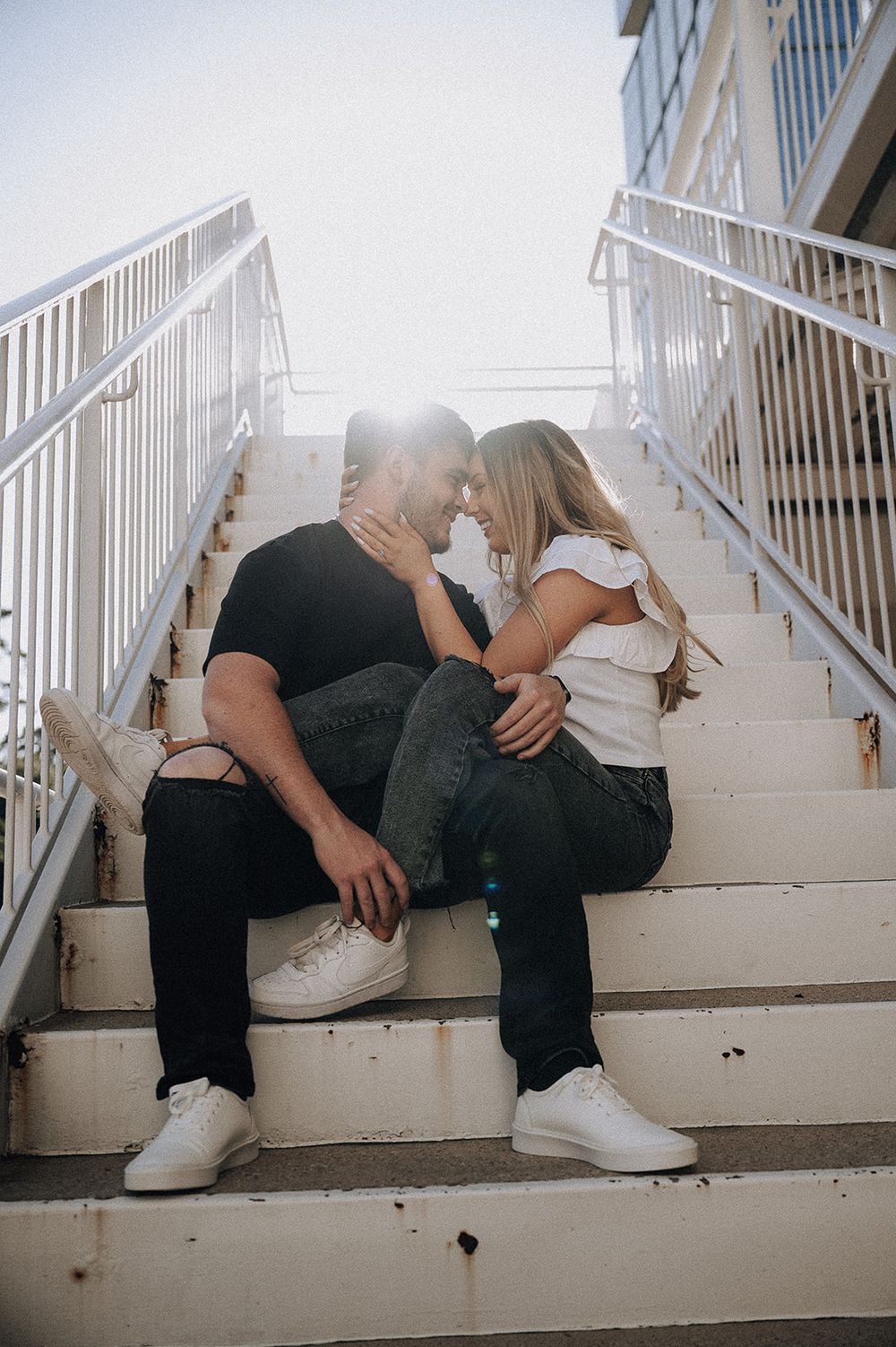 A couple sits intimately on white outdoor stairs with sunlight glowing behind them on a bright sunny day.