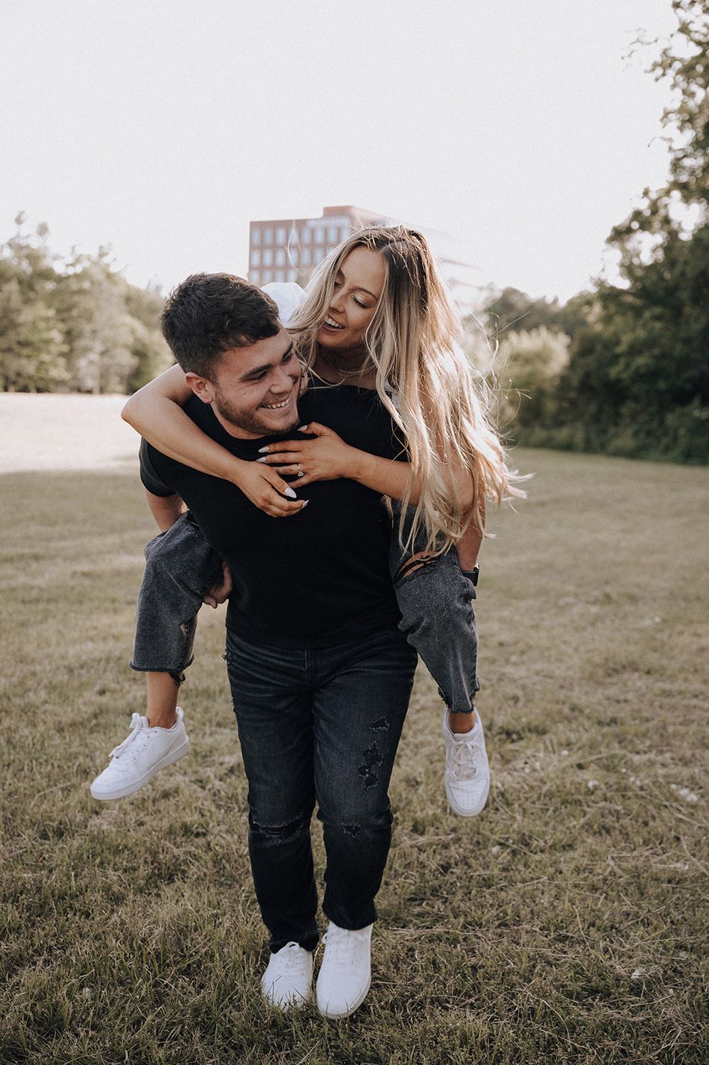 A couple in matching black outfits and white sneakers playfully pose together in a grassy outdoor setting at sunset.