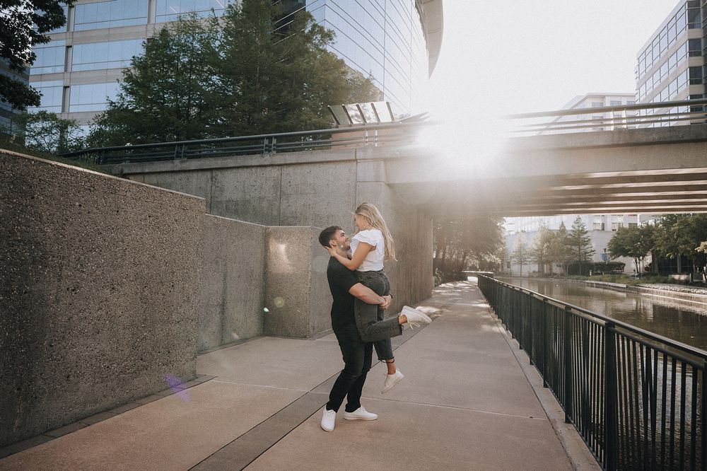 A couple embraces on a waterfront walkway beneath a pedestrian bridge at sunset in an urban setting.
