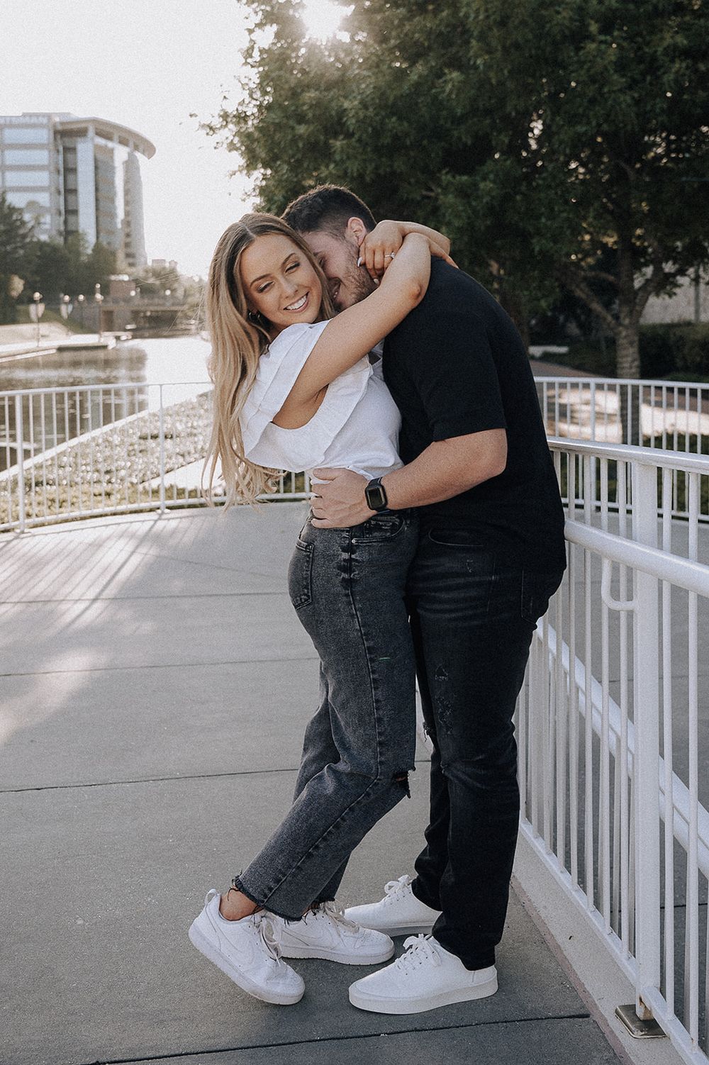A couple embraces on a white railing walkway during golden hour with a city building visible in the background.