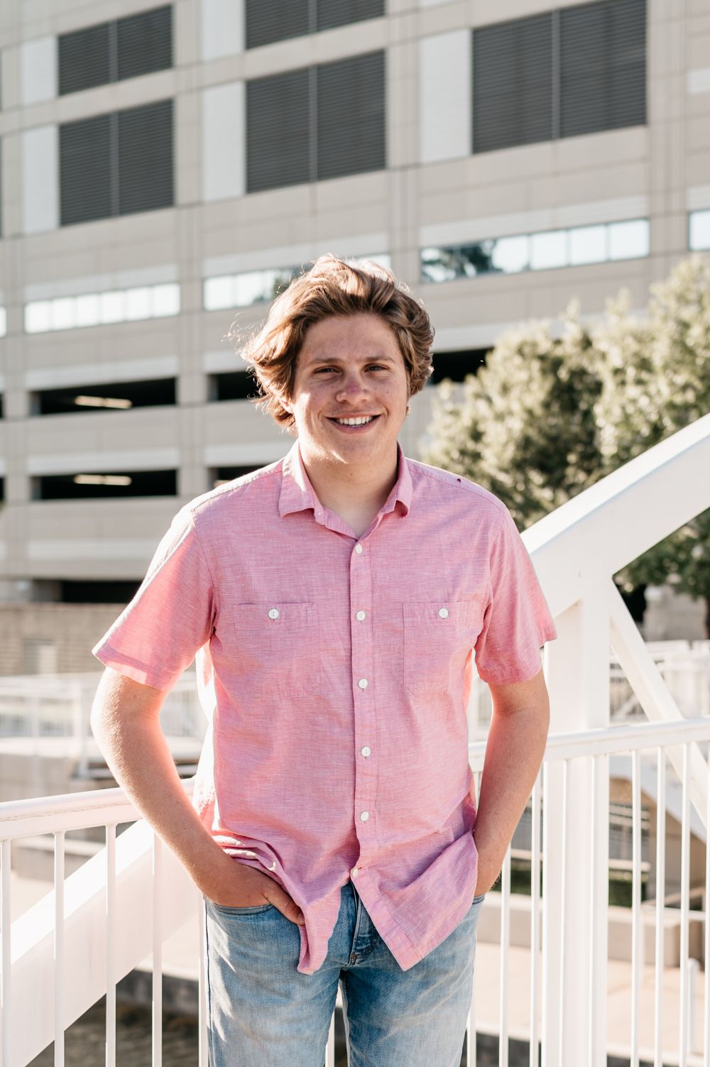 Person wearing pink button-down shirt and jeans stands smiling in front of parking garage on sunny day.