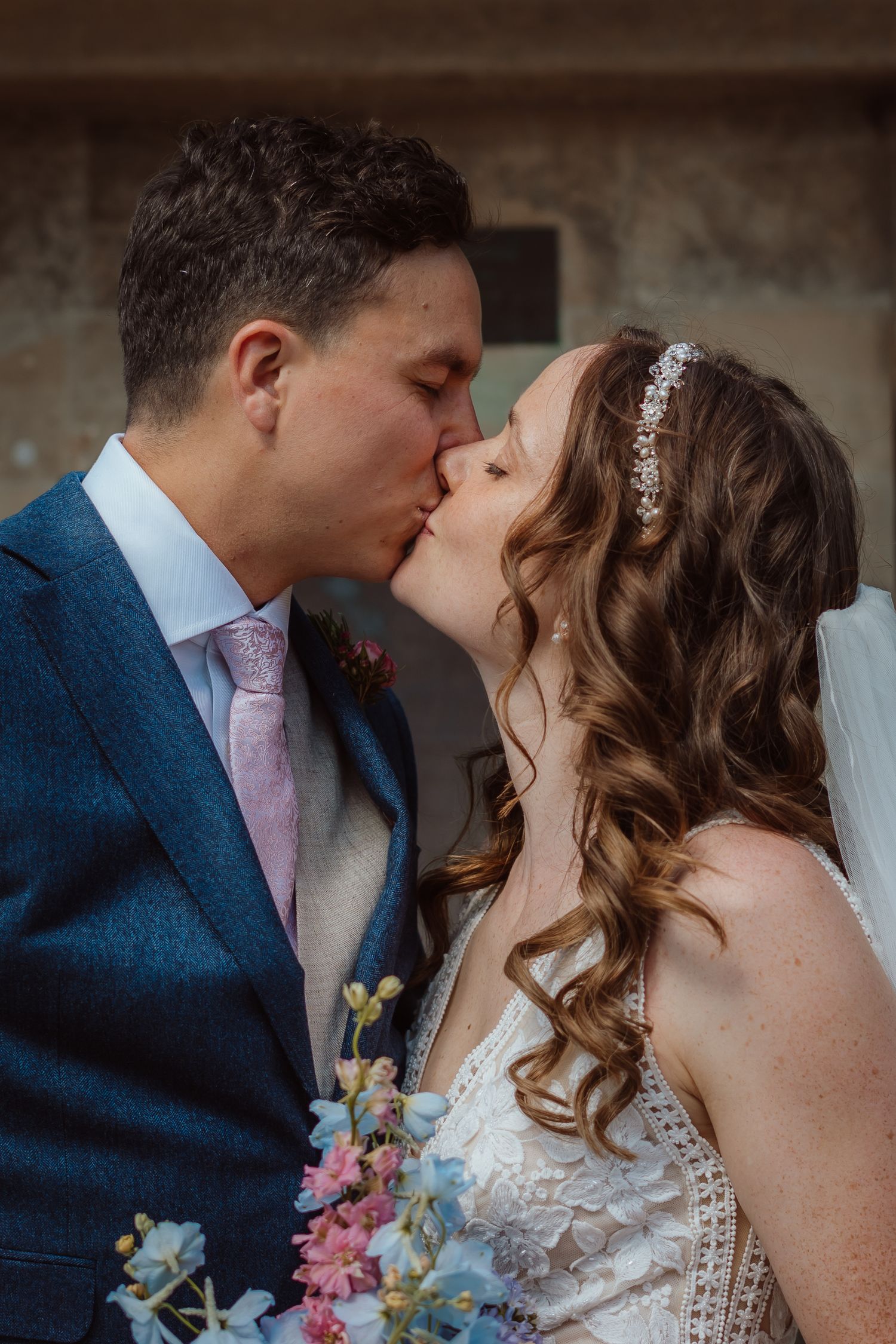 A romantic wedding kiss between newlyweds, with the bride in a lace dress and sparkly hairpiece holding a blue and pink bouquet.