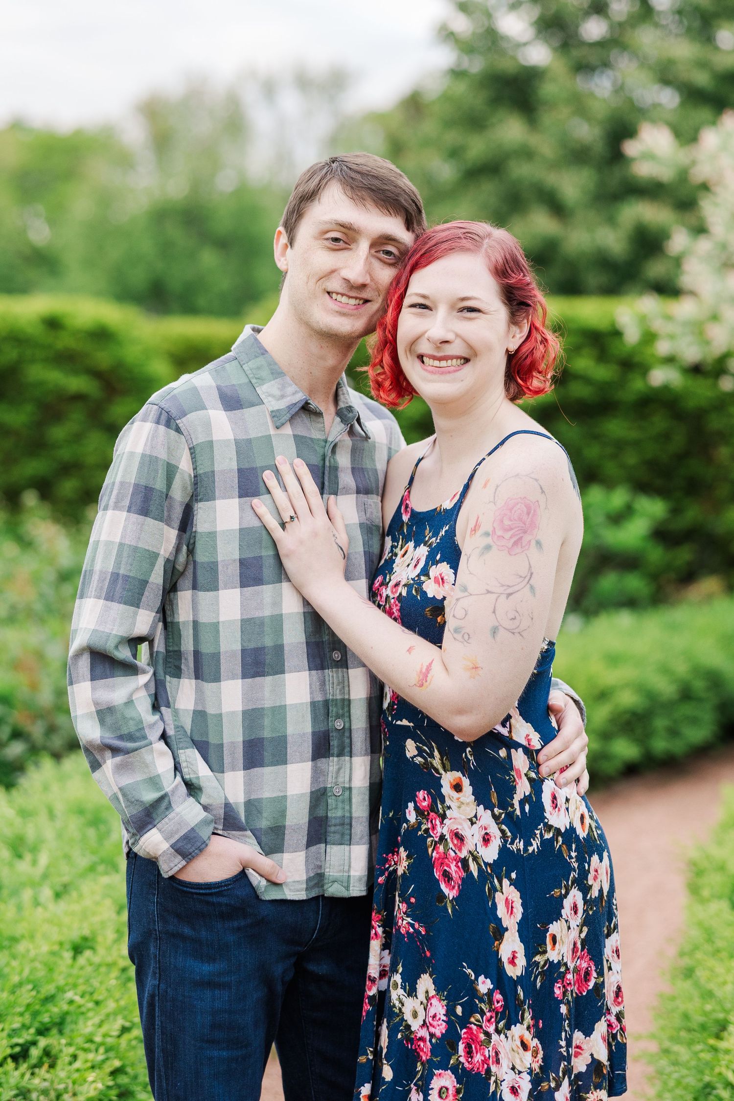 A couple poses outdoors in a garden, wearing a plaid shirt and floral dress against green foliage at Cantigny Park