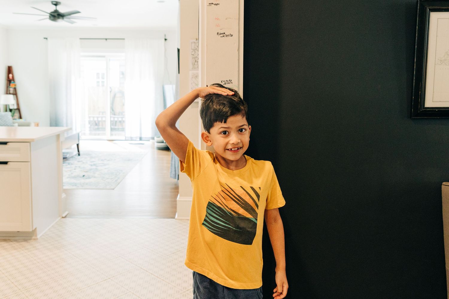 A child in a yellow t-shirt with a green design stands in a bright hallway with dark walls.