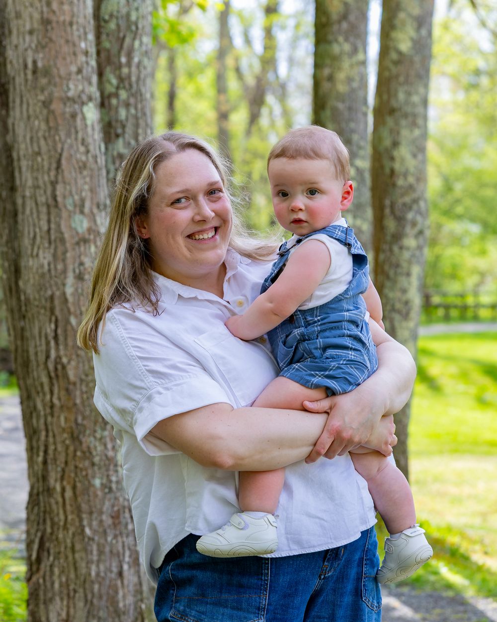 A baby wearing blue overalls and a white shirt is held outdoors by an adult in a white top during a Motherhood photo session in Sudbury, MA