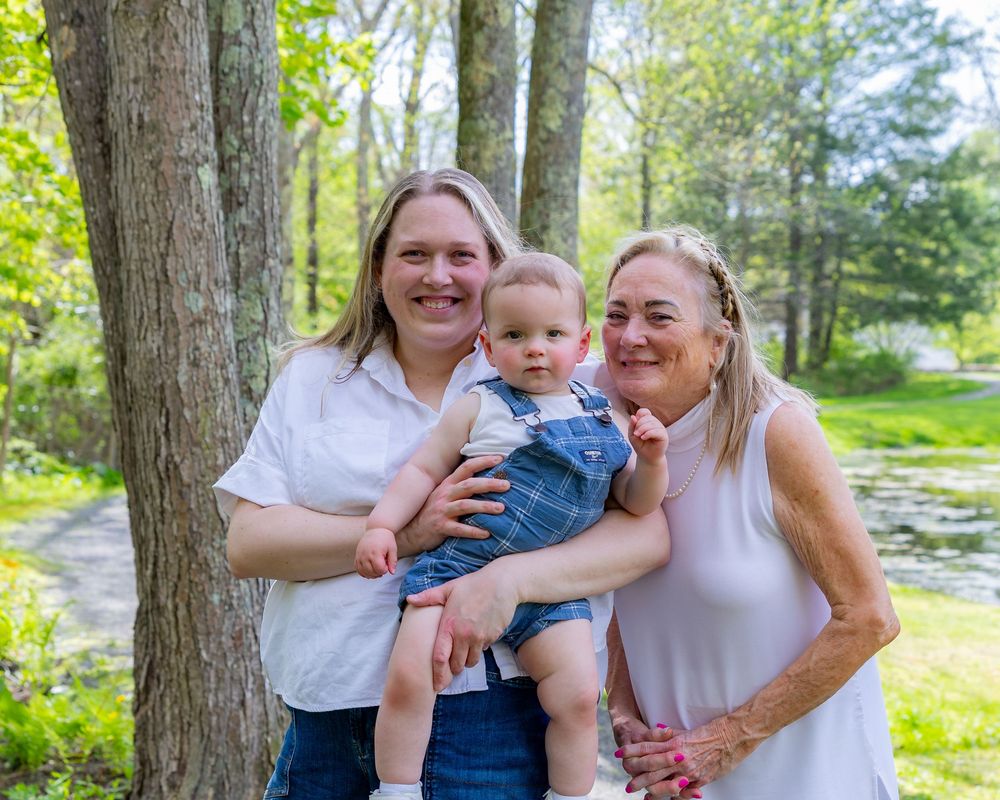 A baby in blue overalls is held by his mother and grandmother during a Motherhood photo session in Sudbury, MA in the Metrowest of Boston.