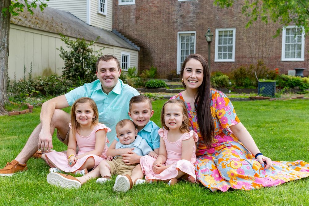 A family group relaxes together on a blanket in their backyard with brick house in background during a family portrait session in Sudbury