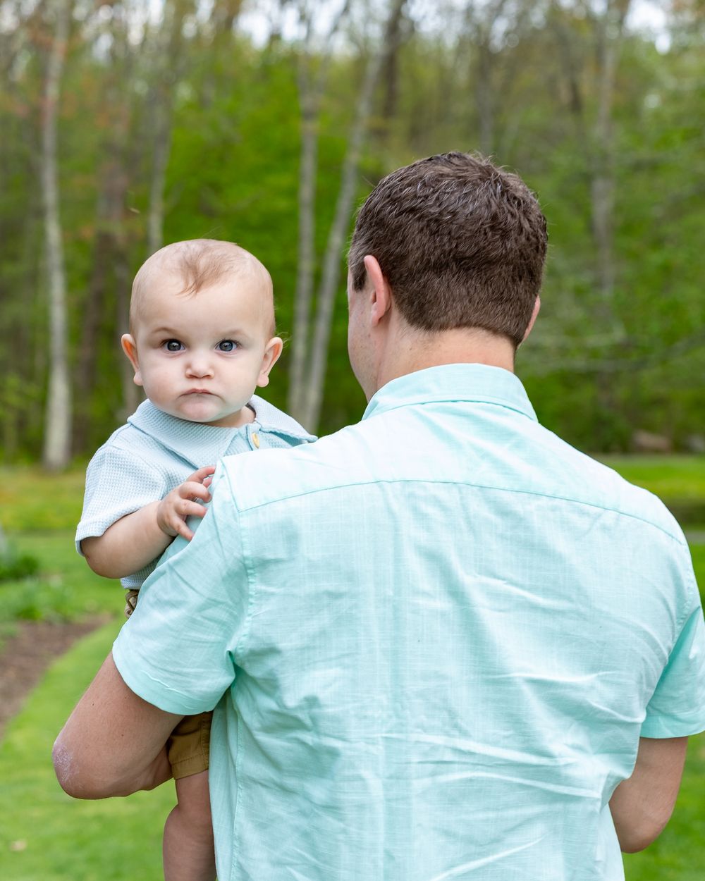 Adult holds small child wearing light blue shirt against wooded natural background during a family portrait session in Sudbury, MA Metrowest