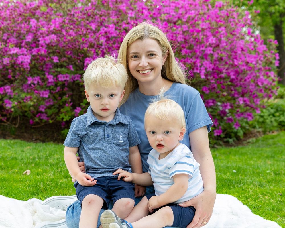A casual outdoor family portrait taken in front of vibrant pink azalea bushes on a sunny day during a family portrait session near Boston.
