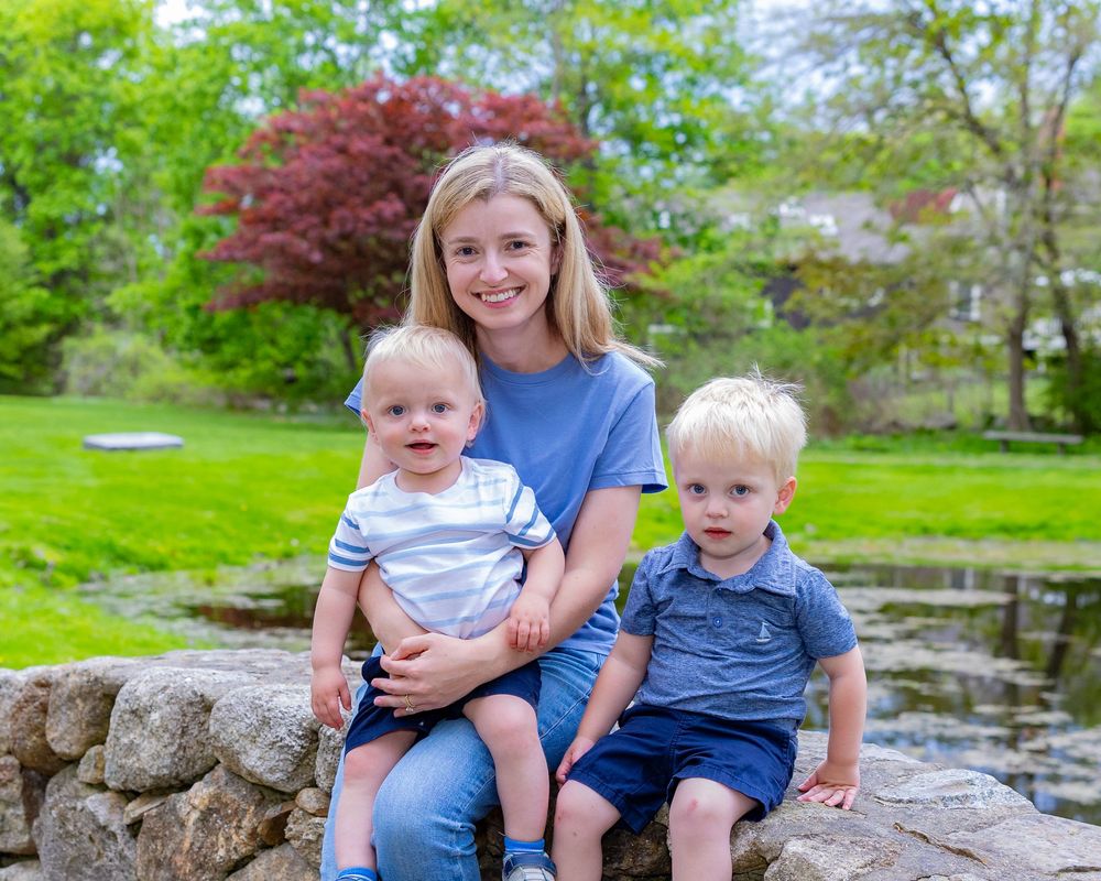 A family portrait captured on stone wall with pond and colorful spring landscaping in background during a family portrait session in Sudbury