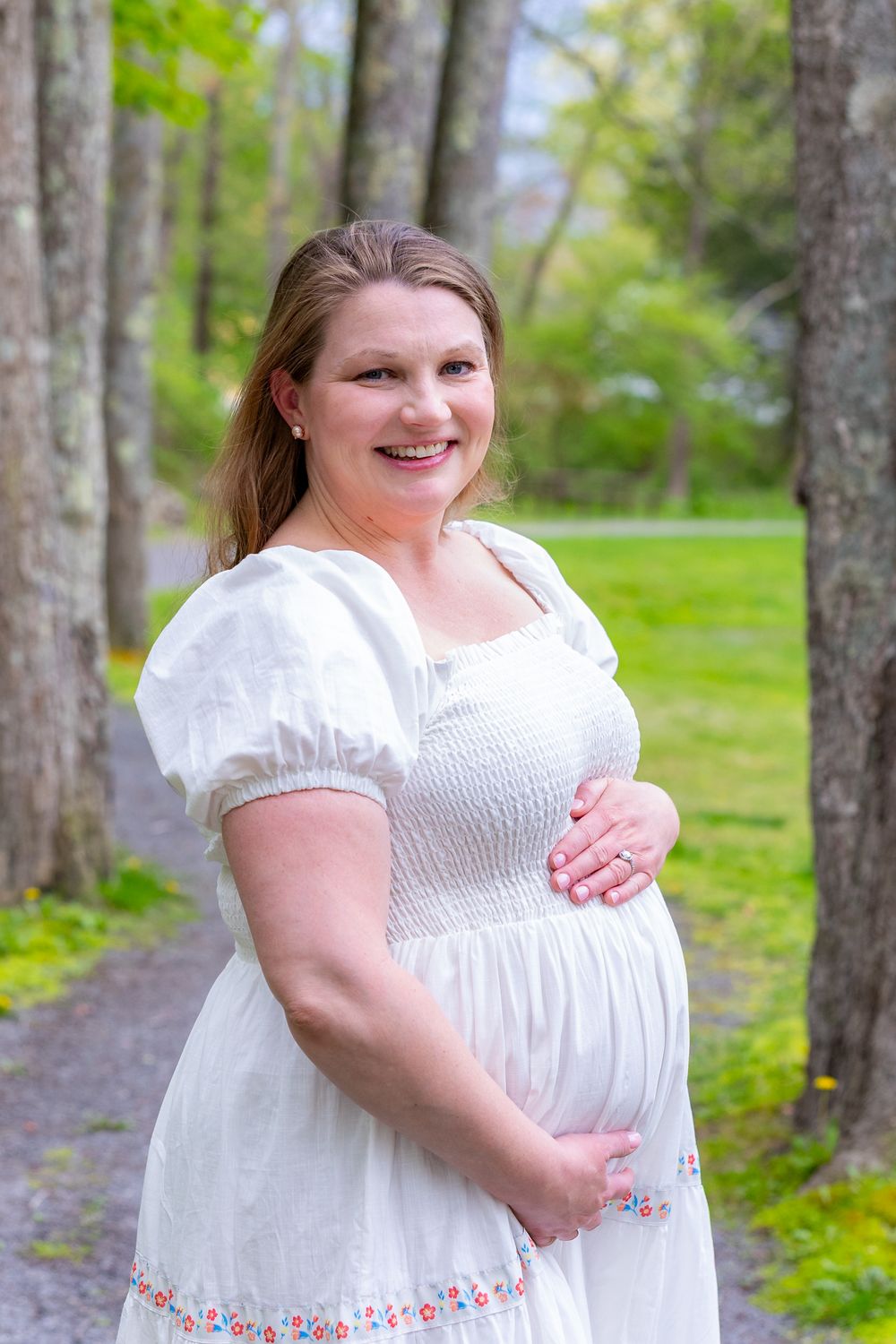 Expectant woman in white dress standing on outdoor path surrounded by spring trees during a family portrait photo session in Sudbury, MA