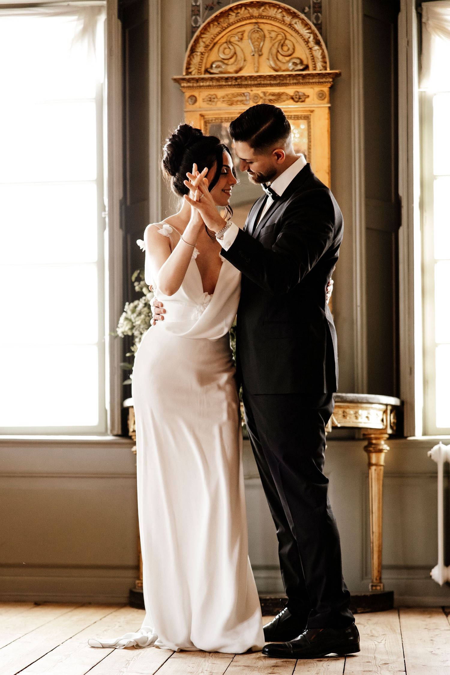 A couple in wedding attire share an intimate moment in an elegant room with ornate gold decor and natural window light.