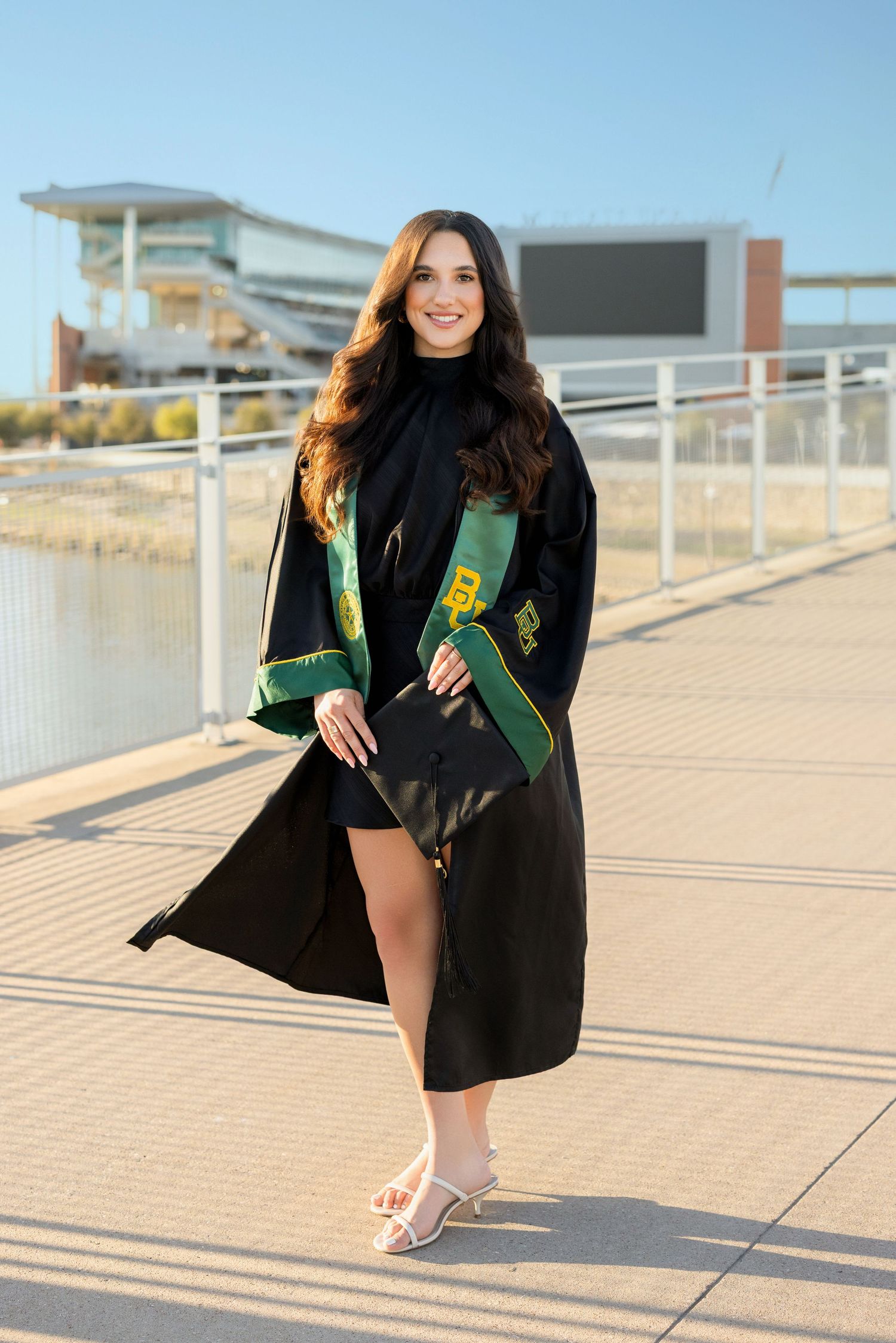 Graduate in cap and gown holding green diploma holder poses on sunny campus walkway near modern building.