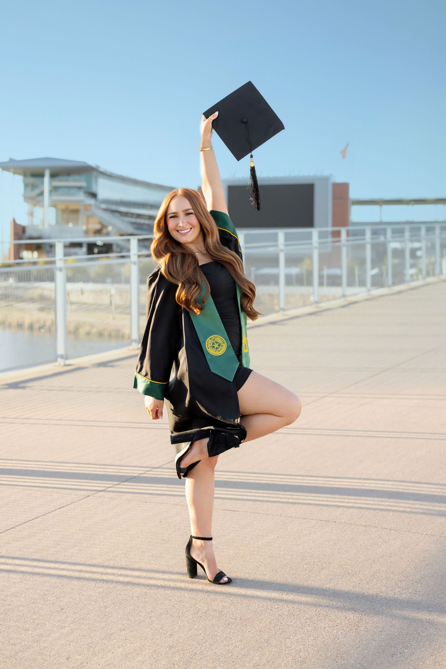 A graduate in cap and gown with sash strikes celebratory pose while holding graduation cap in the air outdoors.