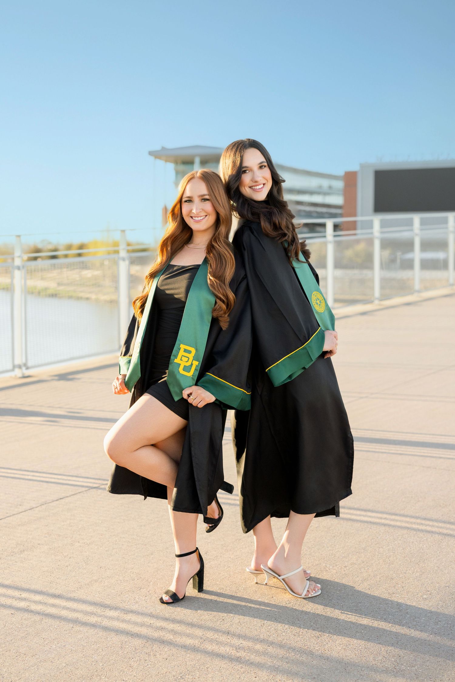 Two graduates in black and green regalia pose together for a celebratory photo on a sunny day at their graduation ceremony.