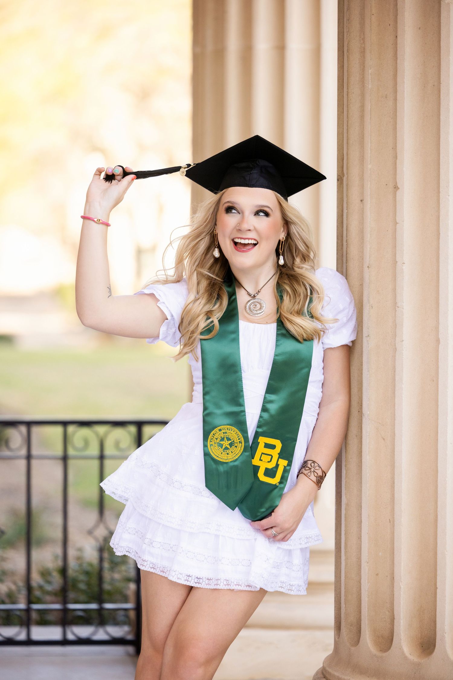 Graduate in white dress and green Baylor University stole playfully poses with graduation cap against stone pillars.