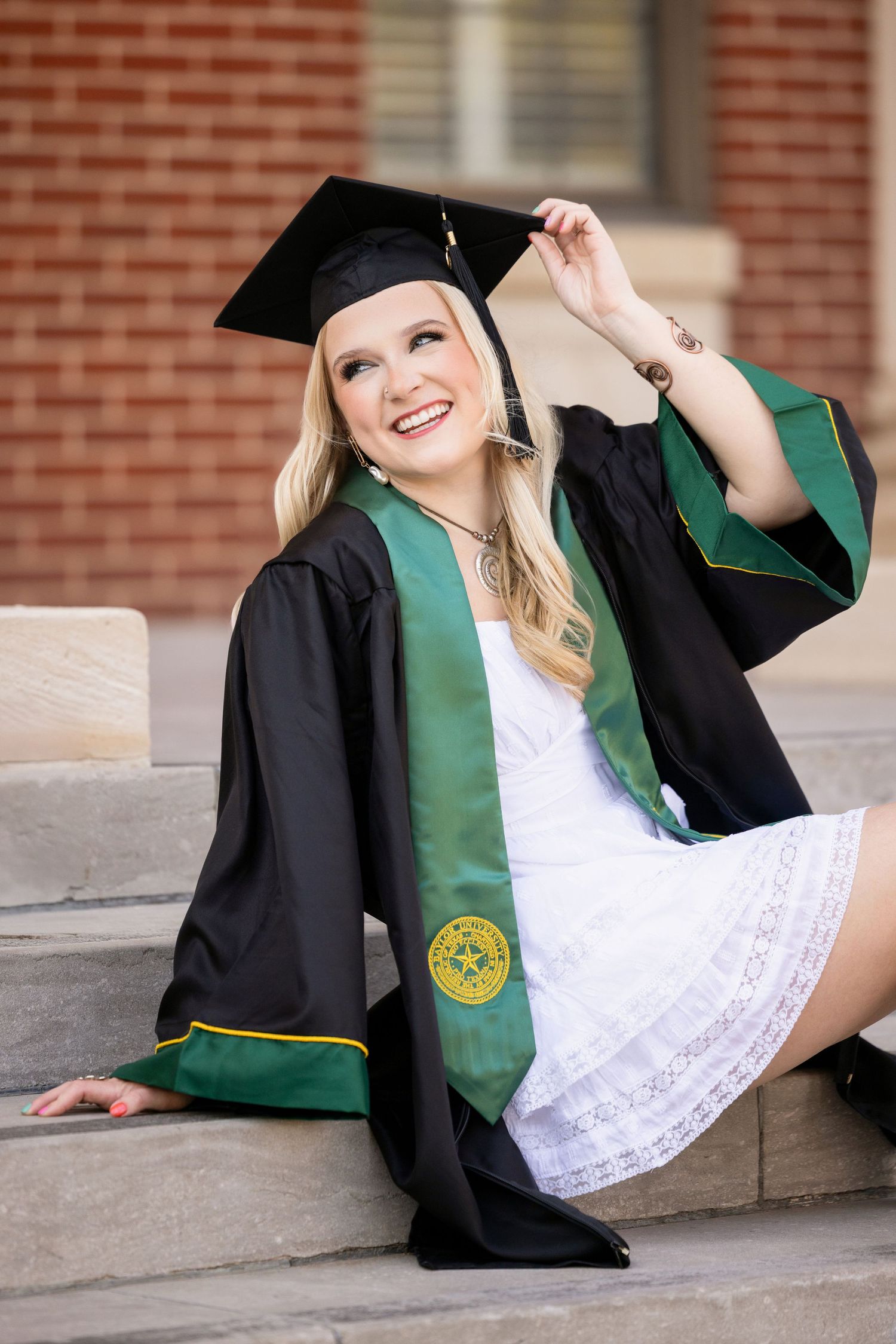 Graduate in black cap and gown with green sash poses on steps of brick building during graduation photo session.