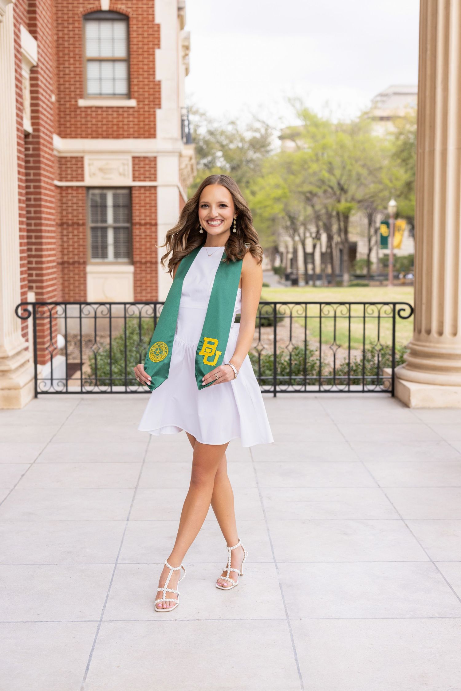 College graduate wearing white dress and graduation stole poses on campus with brick building and columns in background.
