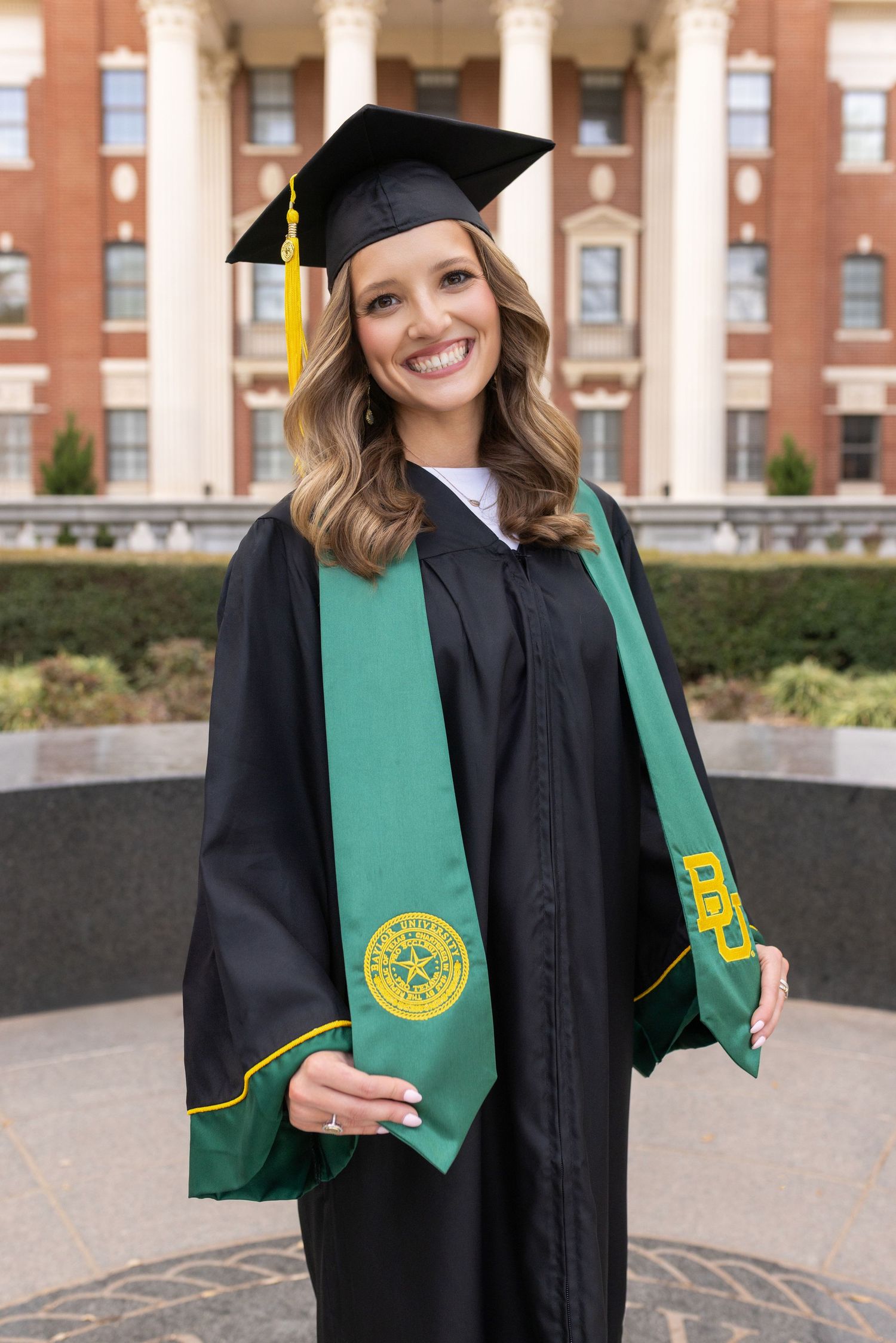 Graduate in black cap and gown with green honors stole poses in front of brick university building.