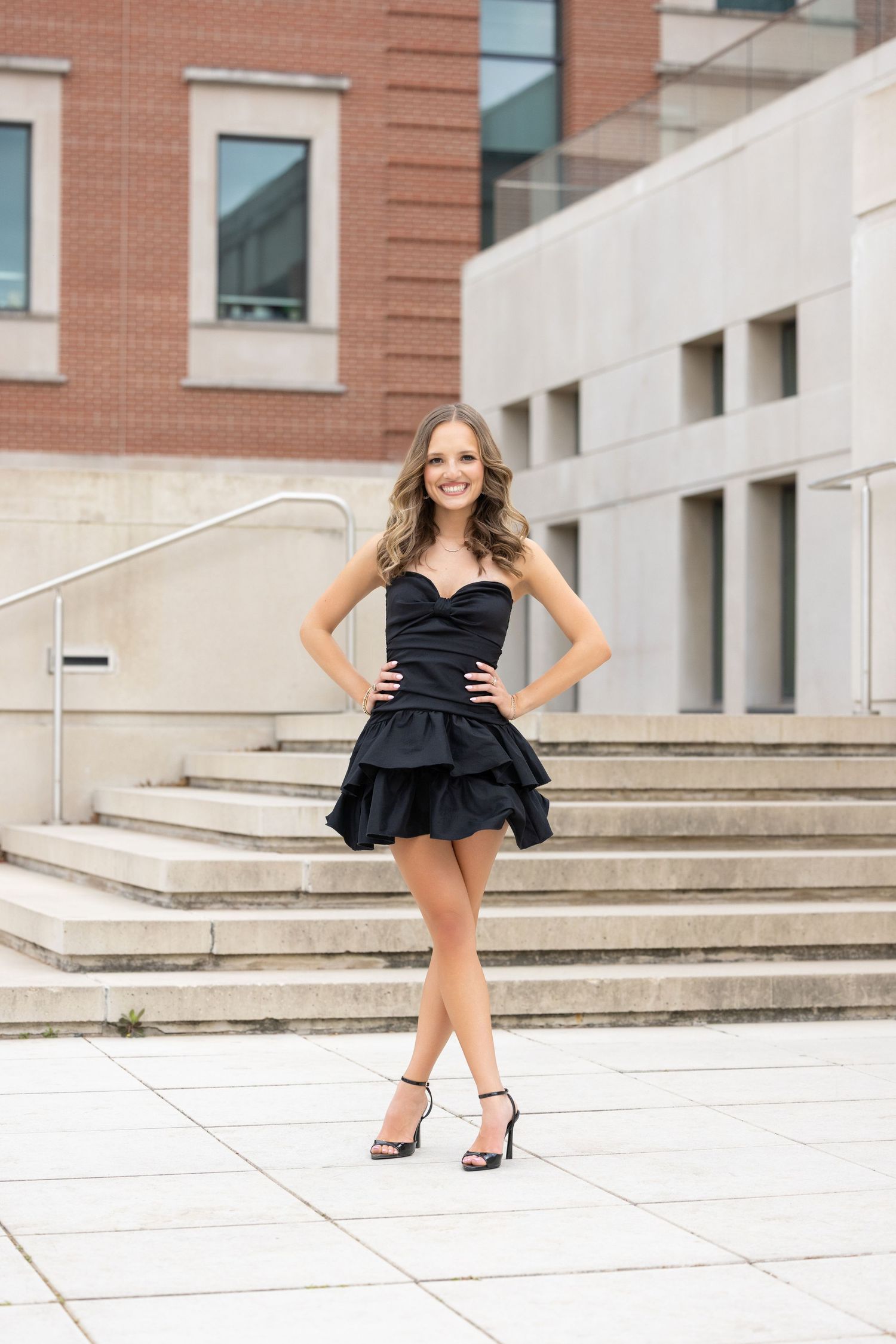 Person in a strapless black cocktail dress with ruffled skirt stands elegantly on outdoor stone steps.