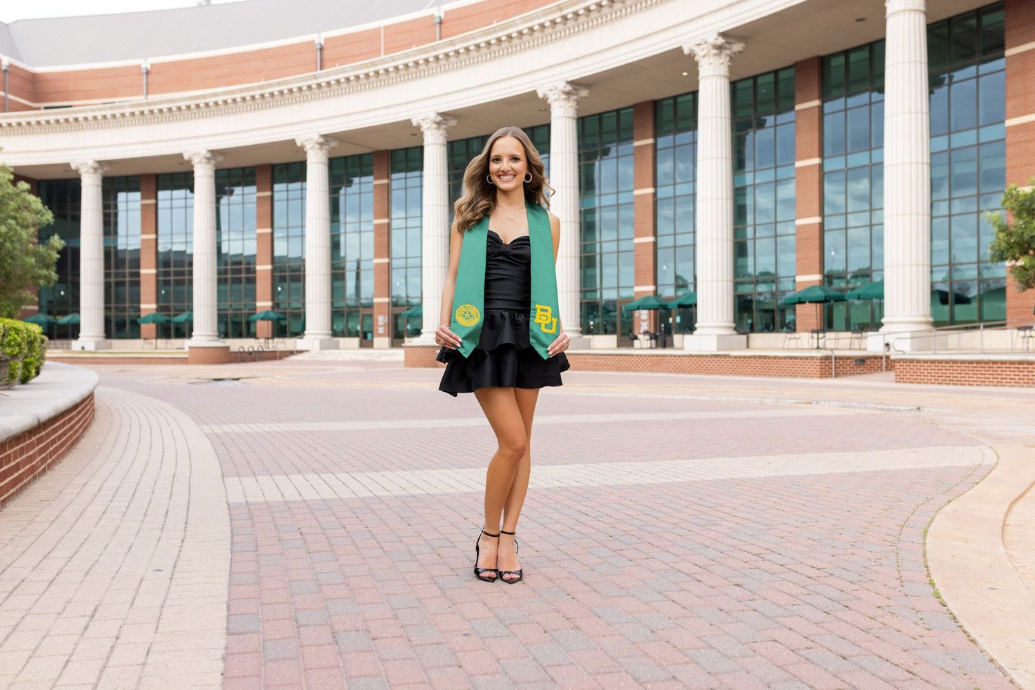 Standing in a green blazer and skirt at a curved brick walkway in front of a large modern building with glass windows.