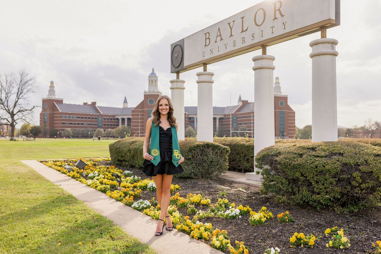 A Baylor University entrance sign with pillars stands above a garden bed of yellow flowers on the campus grounds.
