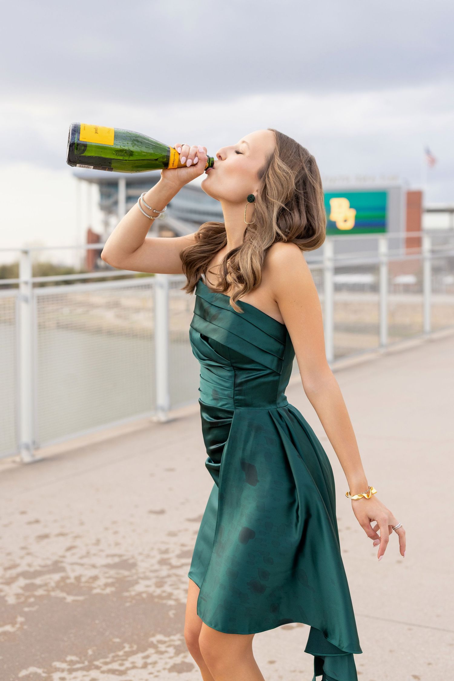 A person in an elegant green cocktail dress drinks from a champagne bottle on an outdoor terrace.