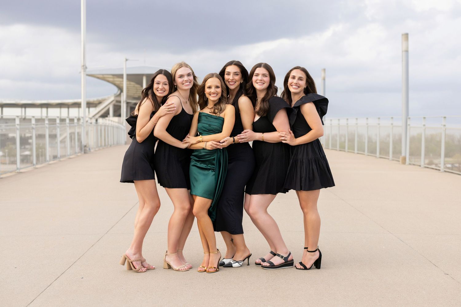 A group of friends in black dresses and one in emerald green pose together on a pier on an overcast day.