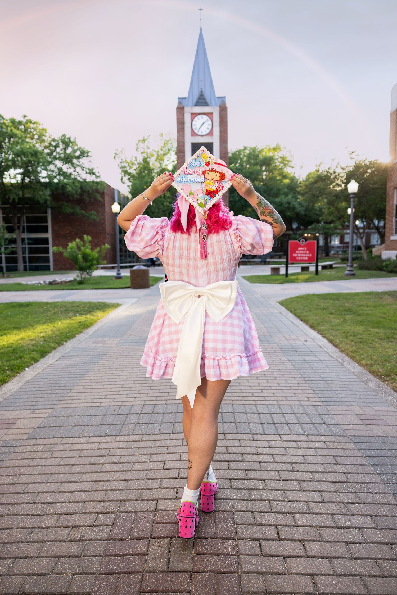 Person in pink gingham dress and sneakers poses playfully on brick path with clock tower in background.
