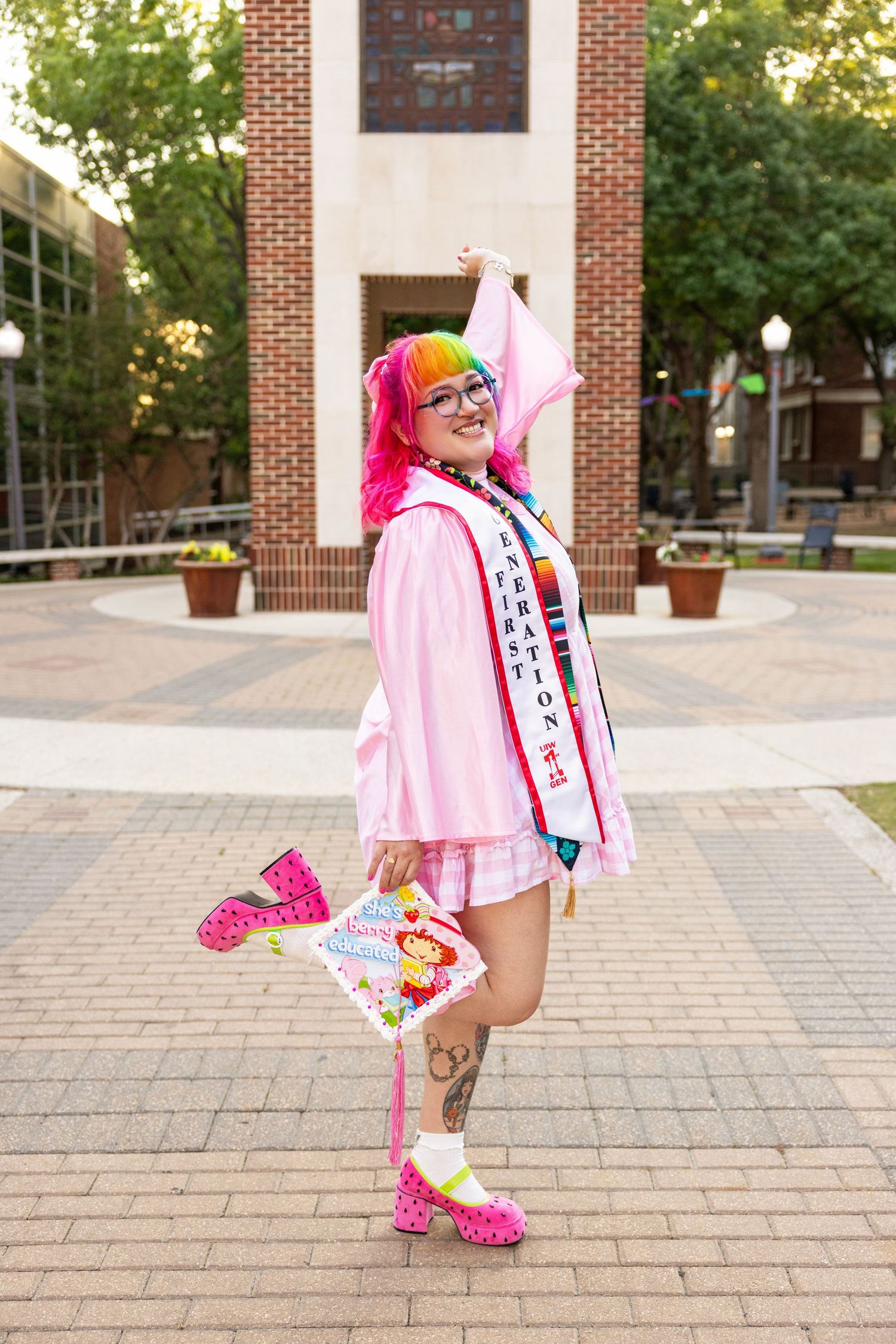 Graduate wearing pink regalia with rainbow hair and sparkly shoes poses playfully on a brick courtyard.