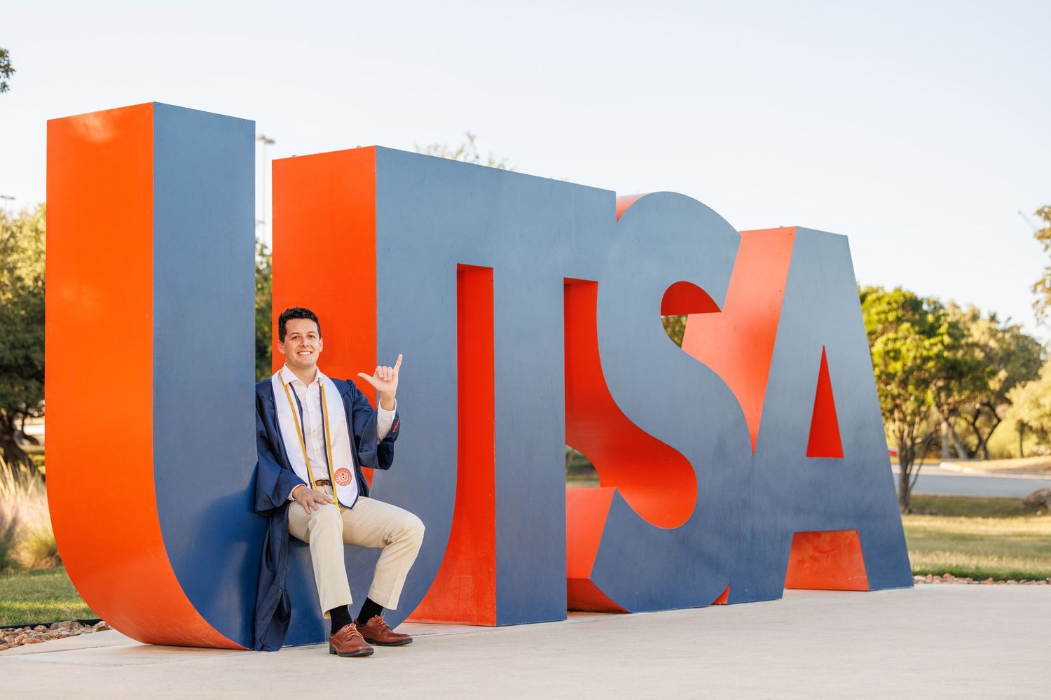 Large orange and gray "UTSA" letters form an outdoor sign installation with a person sitting casually on the first letter.
