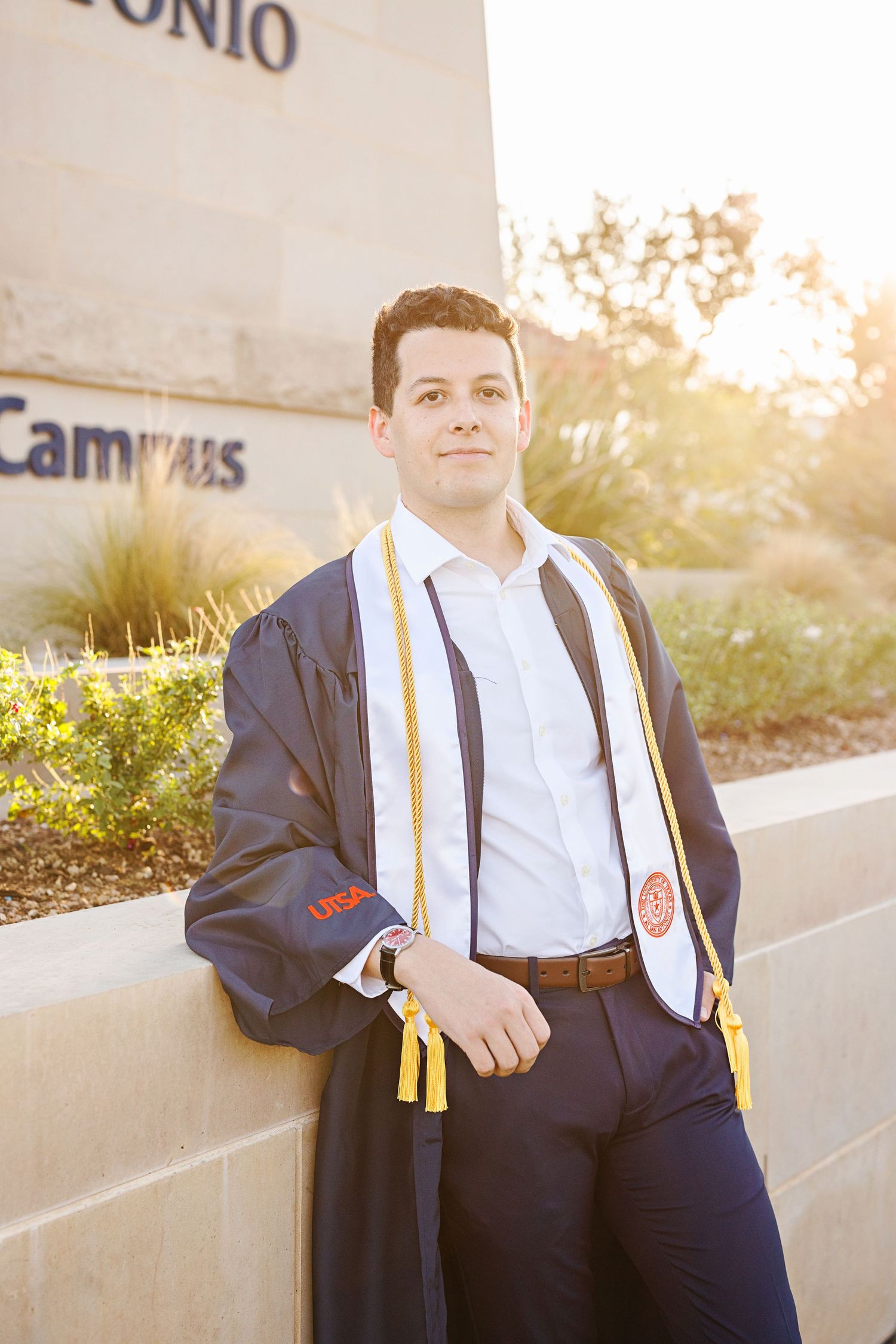 Graduate in cap and gown stands outdoors during golden hour wearing academic regalia and honor cords.