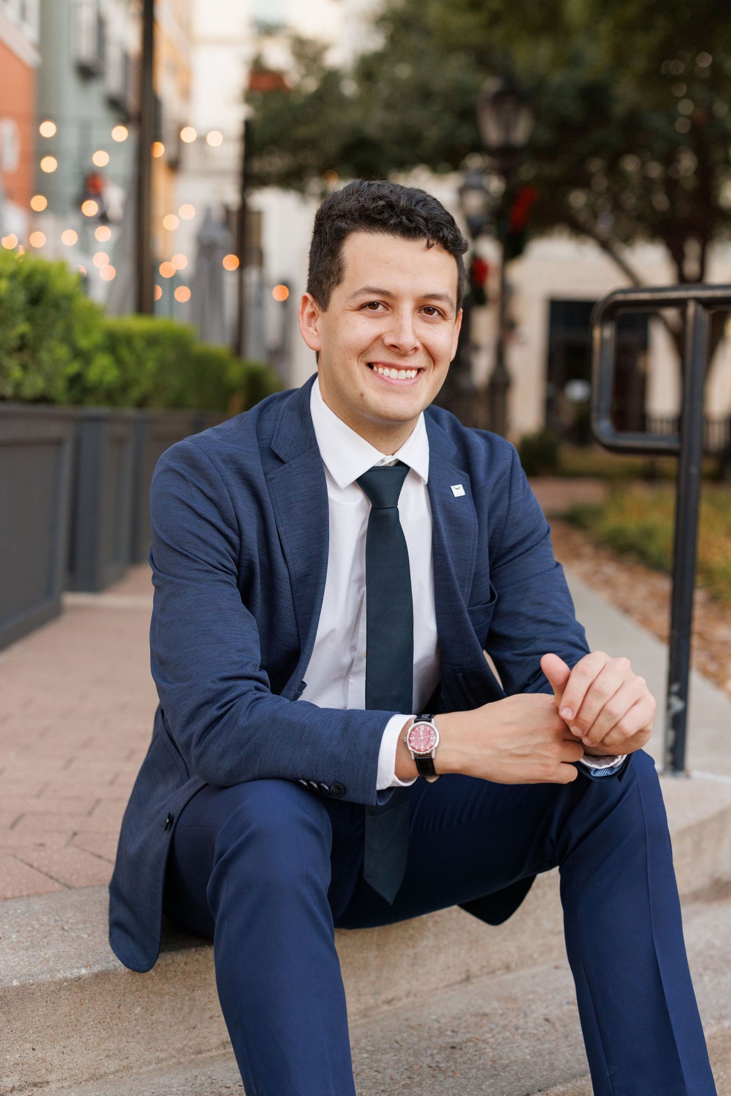 Professional portrait of someone in a navy suit sitting casually on concrete steps in an urban setting.