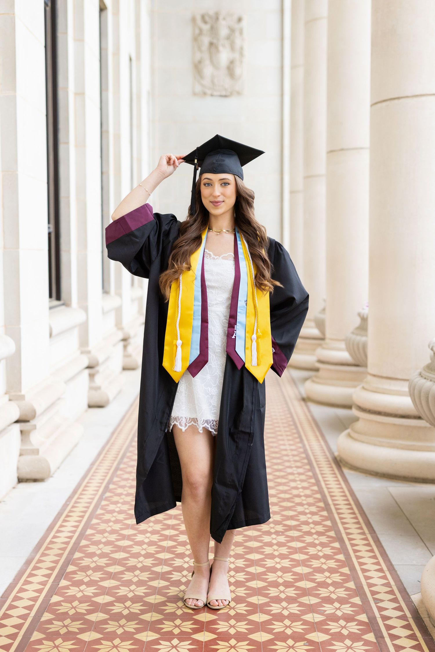 Graduate in cap and gown with gold honors stole walks through elegant columned hallway with tiled floor pattern.