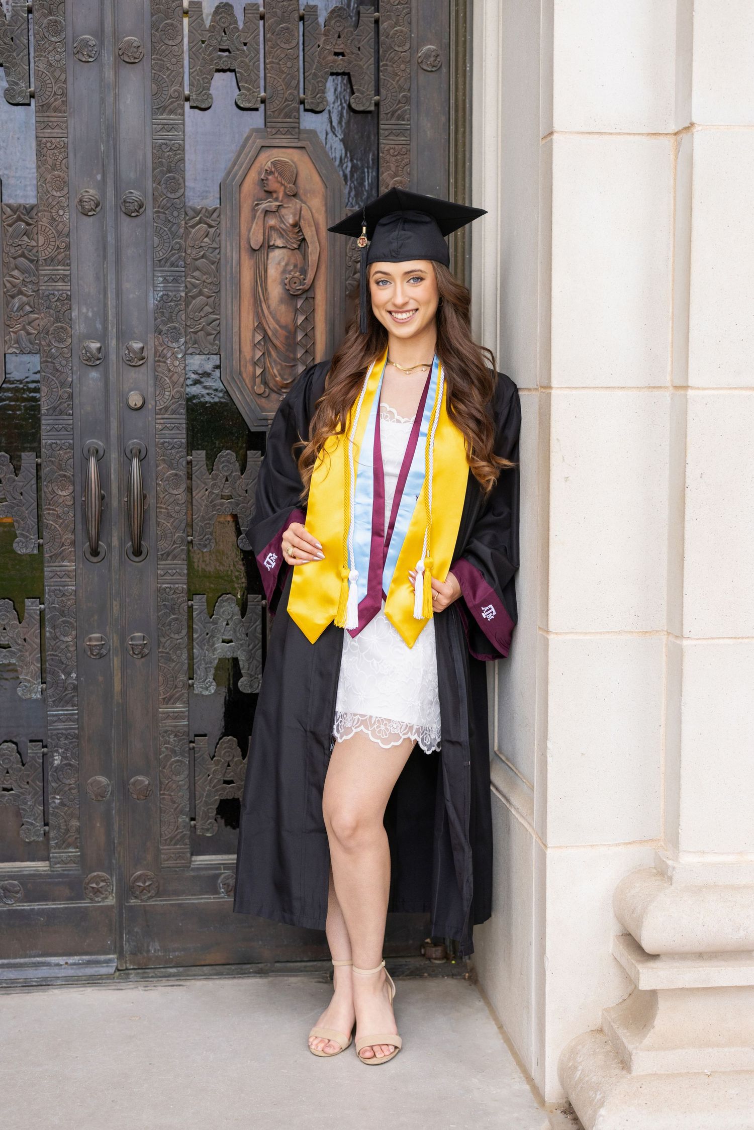 Graduate in cap and gown with gold honors stole stands by ornate church door in white dress and sandals.