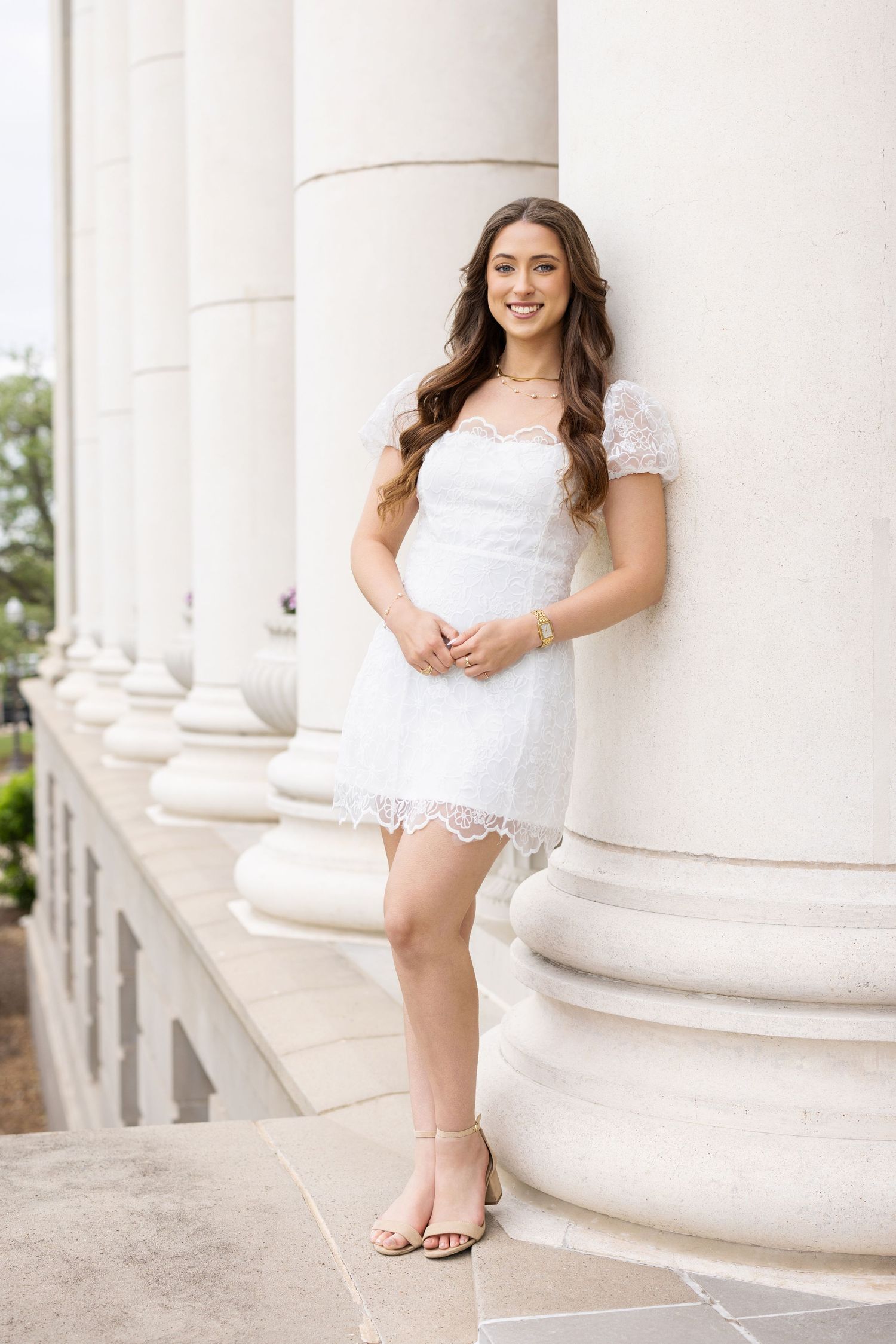 Young woman in white lace dress with short sleeves poses elegantly against classic stone columns on a balcony.