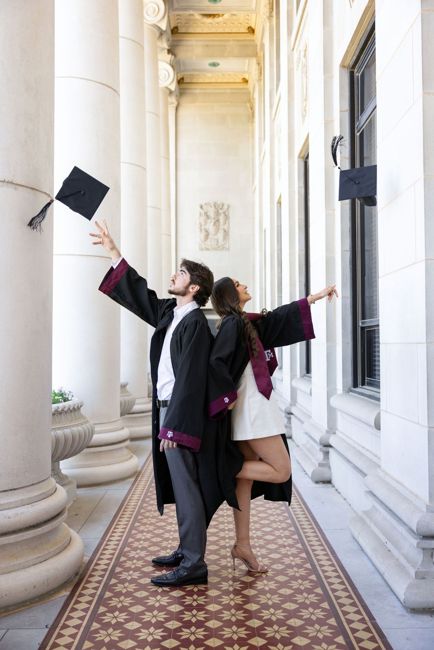 Two graduates celebrate in black graduation gowns while posing along a classical colonnade with white pillars.