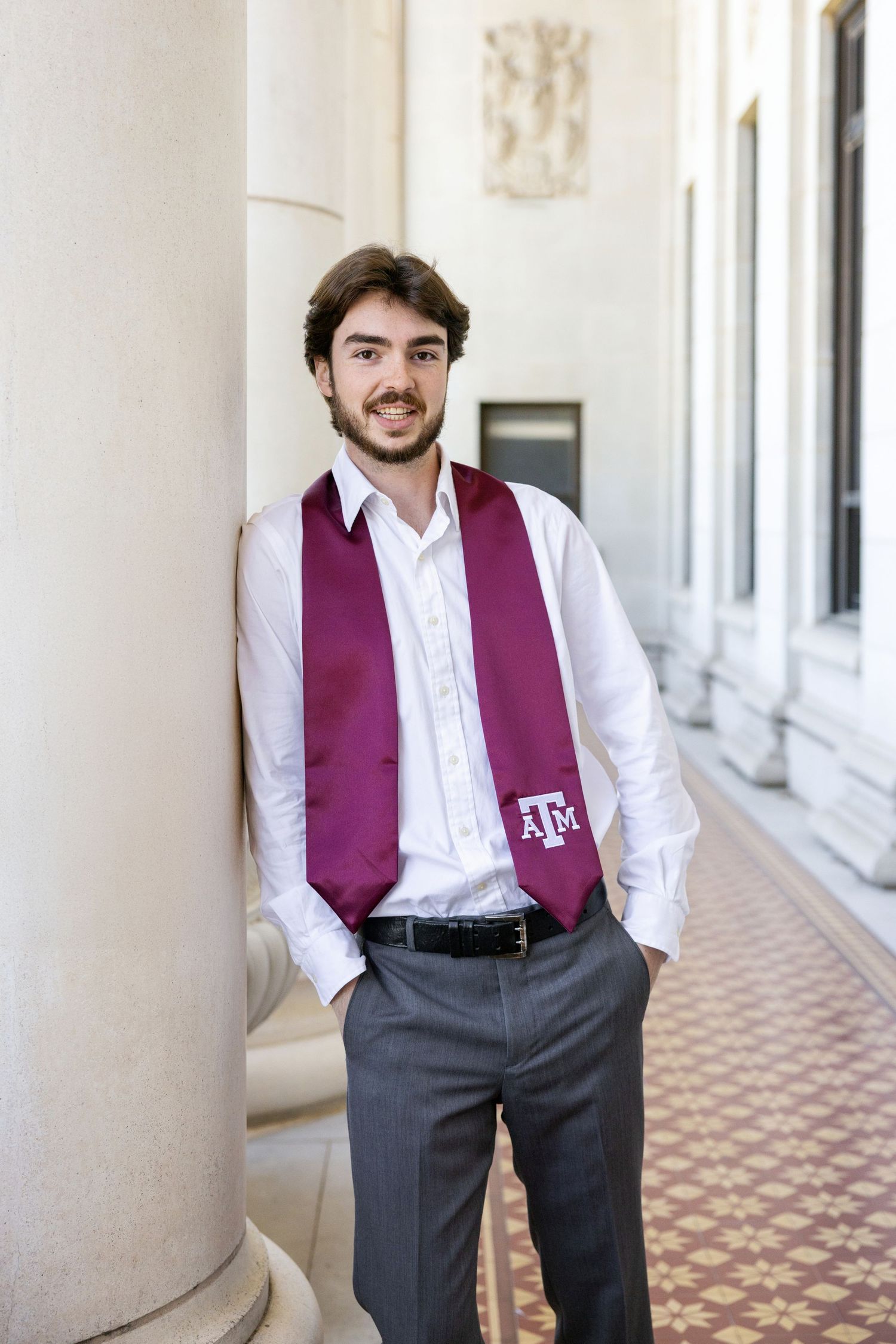 Graduate wearing a purple Texas A&M stole with white dress shirt and gray slacks poses in an ornate hallway.