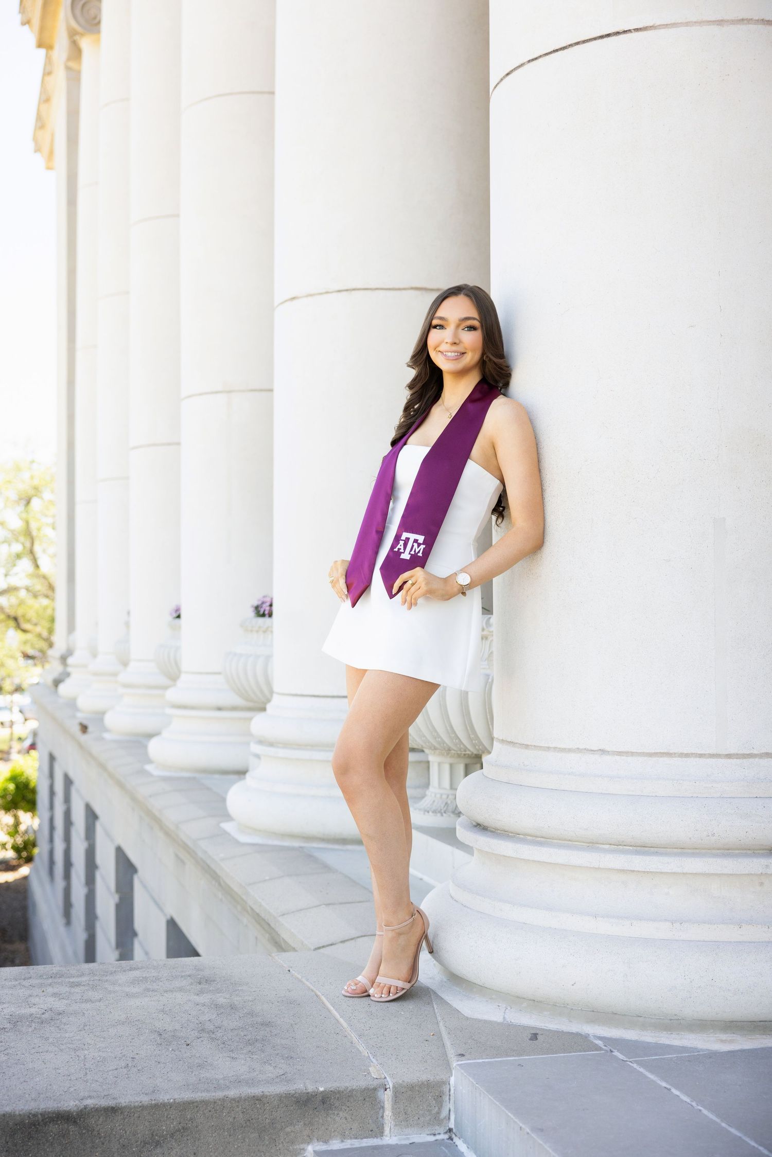 Graduation photo at a columned building with white dress, maroon stole, and heels on stone steps.