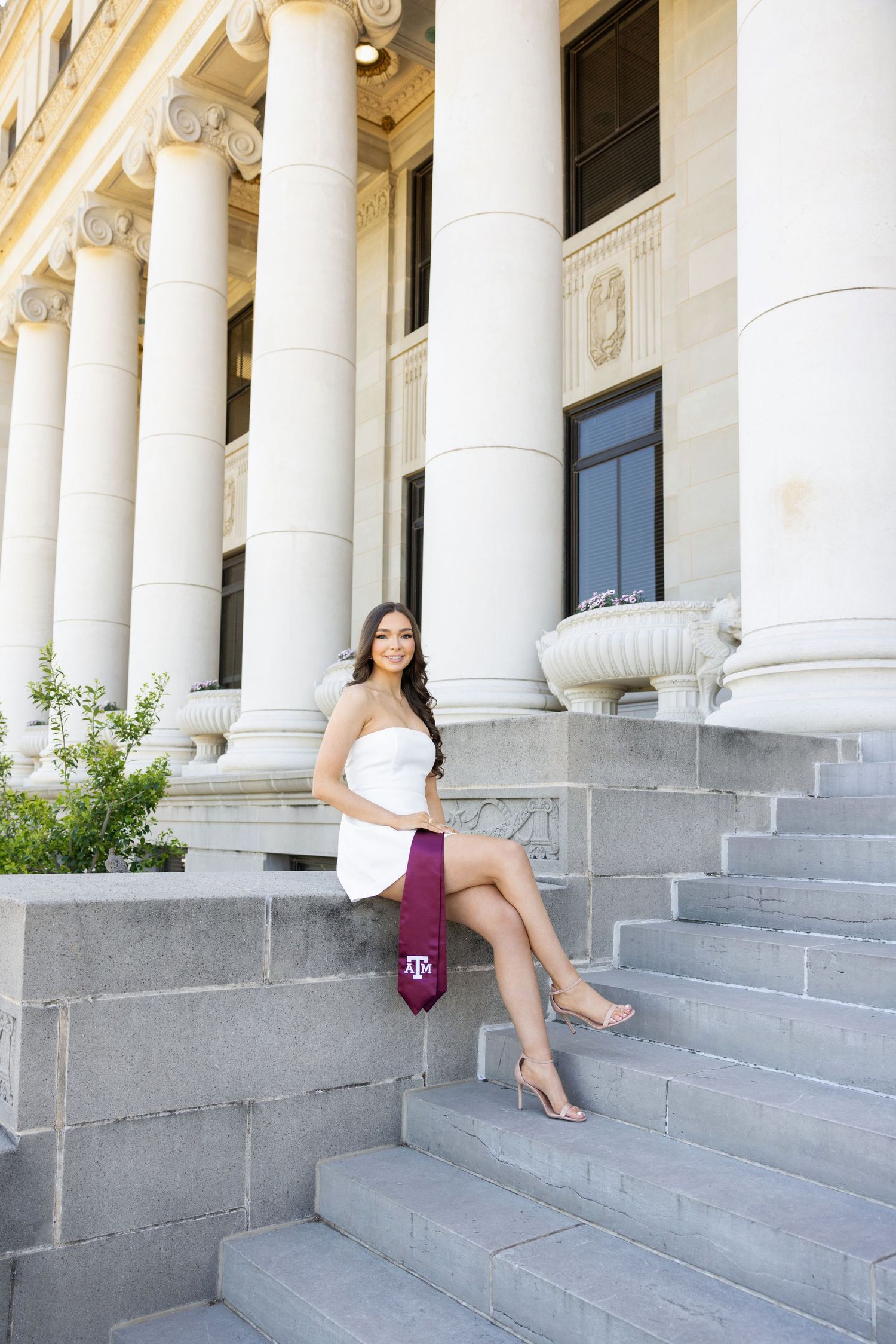 Graduate in white dress sits on stone steps of grand building with classical columns at Texas A&M.
