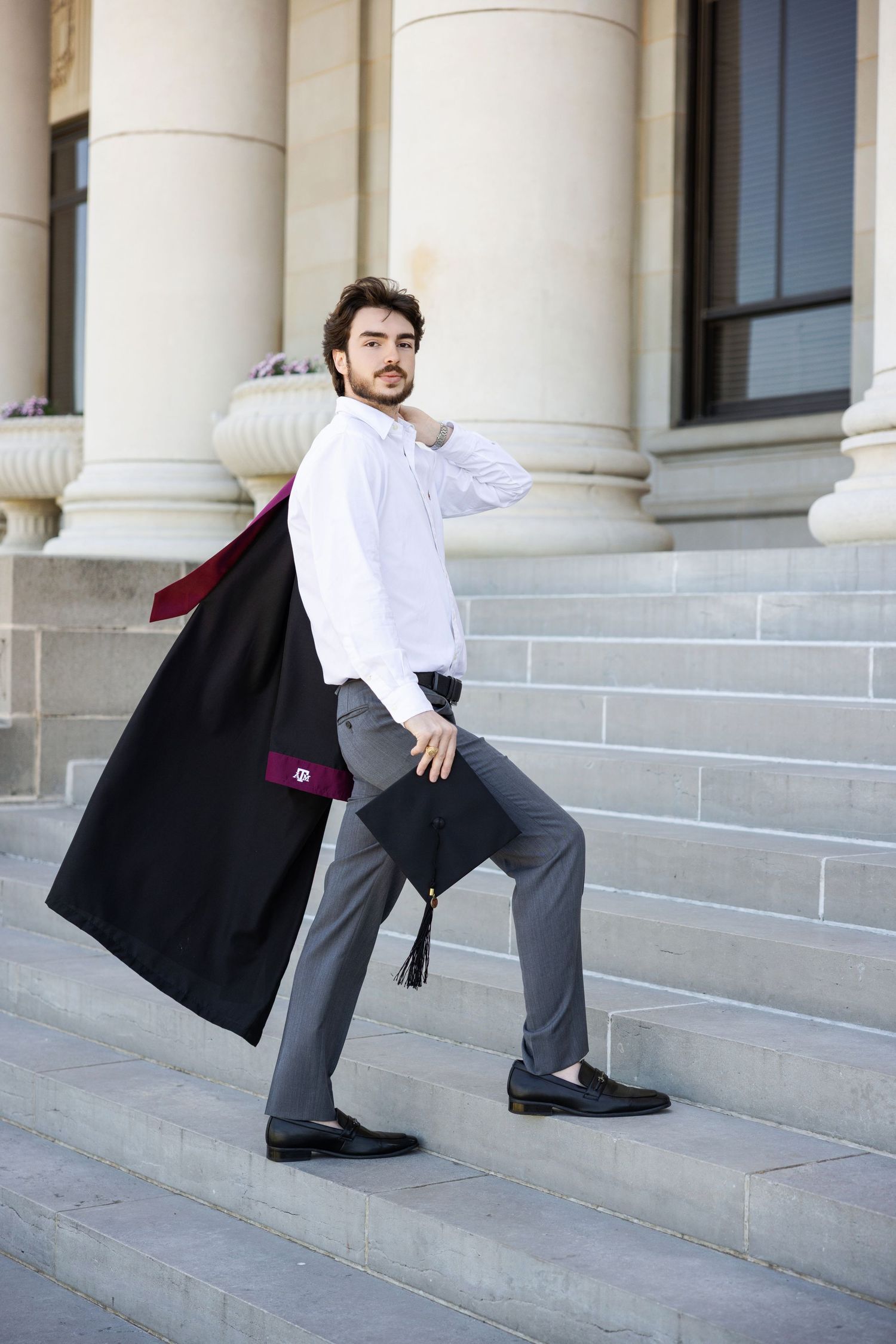 Professional wearing a white dress shirt, gray slacks and black overcoat stands on stone steps near classical columns.