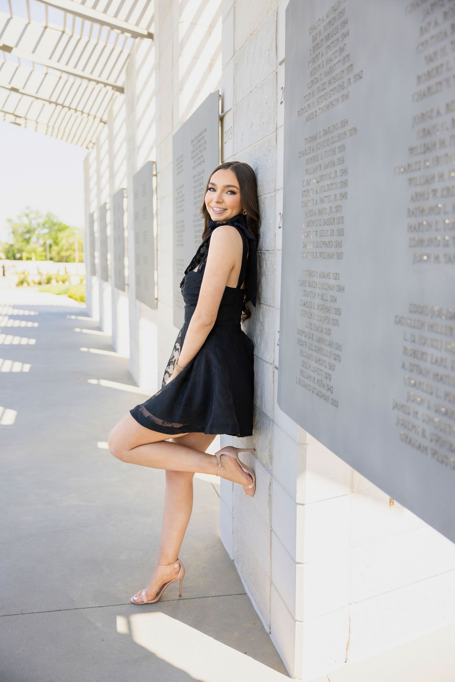 A figure in a black sleeveless dress poses playfully against a white columned wall on a sunny day.